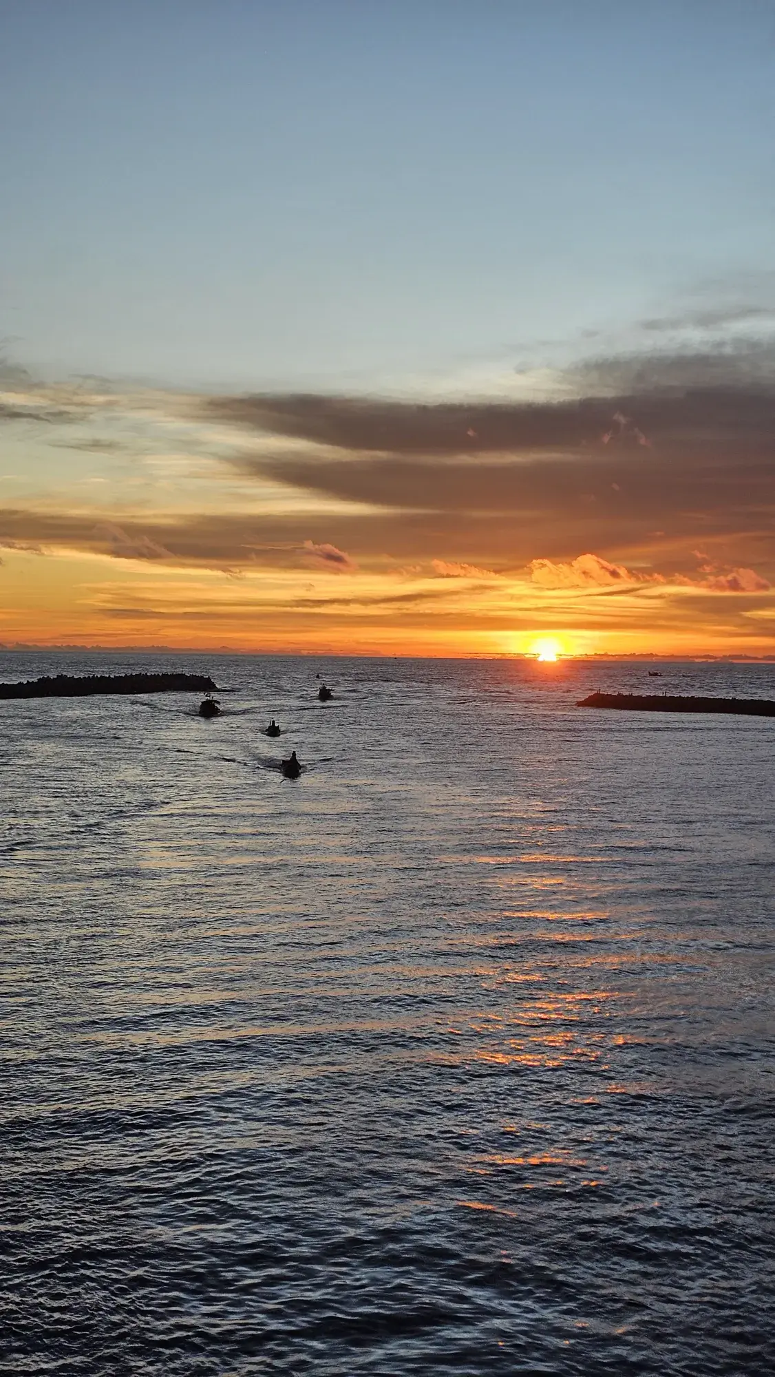 Four small boats heading out past the breakwaters at Azheekal at sunset, the sun low over open sea